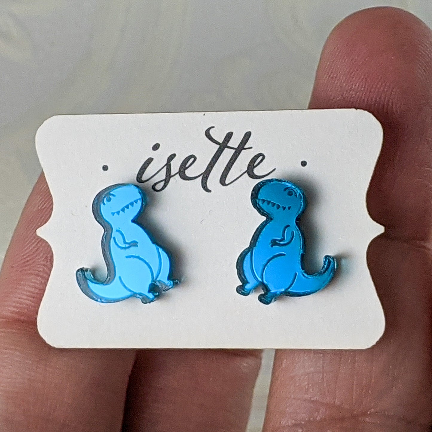 Cartoon Tyrannosaurus Rex stud earrings in teal mirrored acrylic.  The dinosaurs face towards each other and are mounted on an earring card.  By Isette.