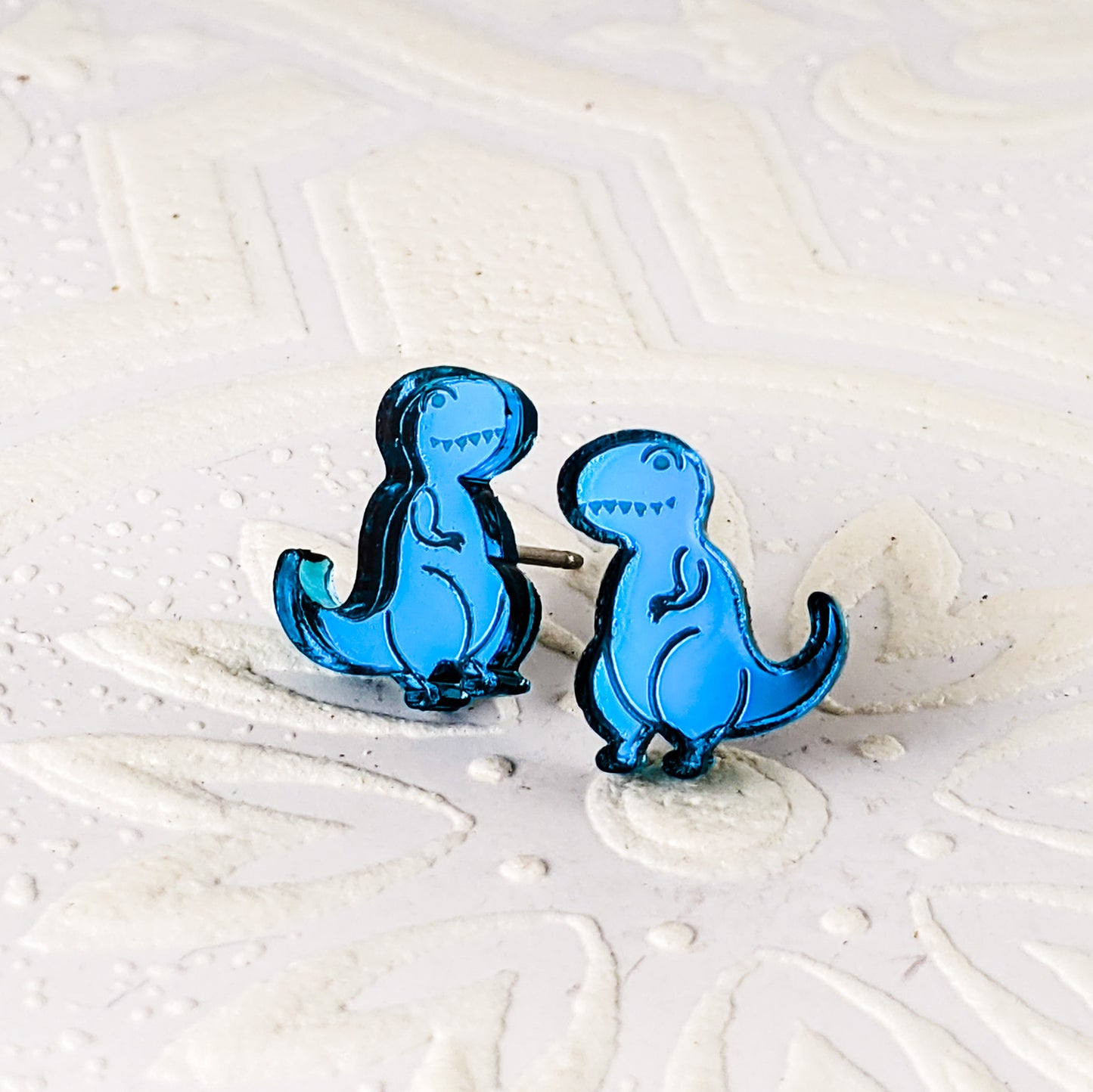 Cartoon Tyrannosaurus Rex stud earrings in teal mirrored acrylic.  The dinosaurs are mirrored, and face towards each other.  By Isette. 