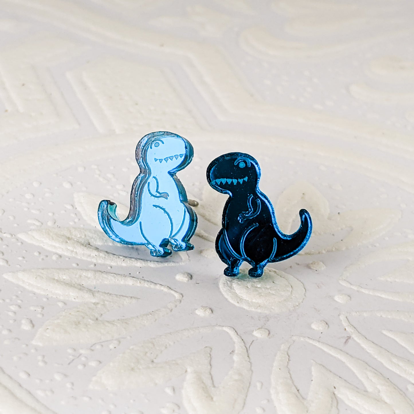 Cartoon Tyrannosaurus Rex stud earrings in teal mirrored acrylic, photographed to show the shine of the mirrored acrylic.  The dinosaurs are mirrored, and face towards each other.  By Isette.