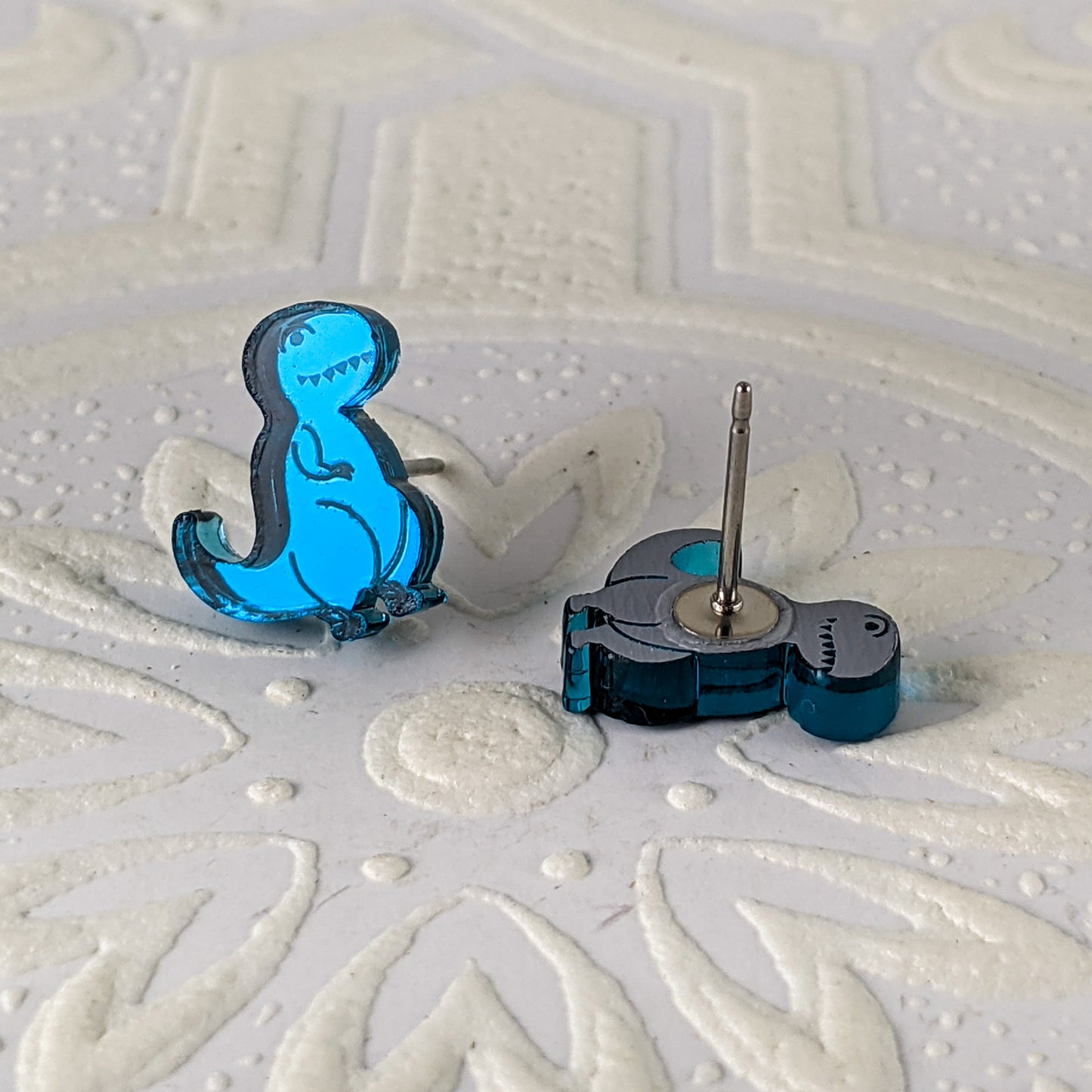Cartoon Tyrannosaurus Rex stud earrings in teal mirrored acrylic.  One Tres is laying down, showing the stainless steel earring post.  By Isette.