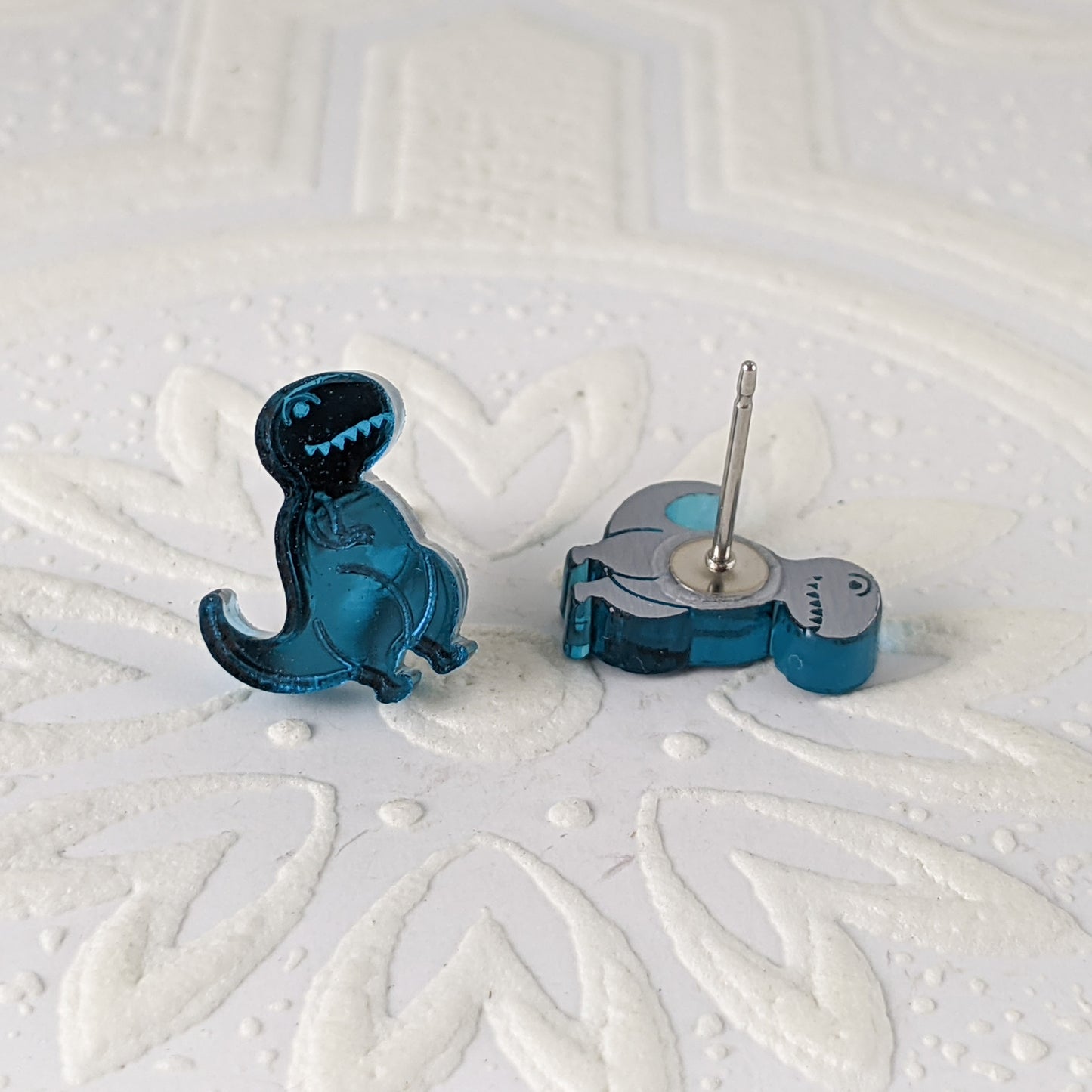 Cartoon Tyrannosaurus Rex stud earrings in teal mirrored acrylic.  Photograpehd so the shine is visible, and one Trex is face down to show the stainless steel post. By Isette.