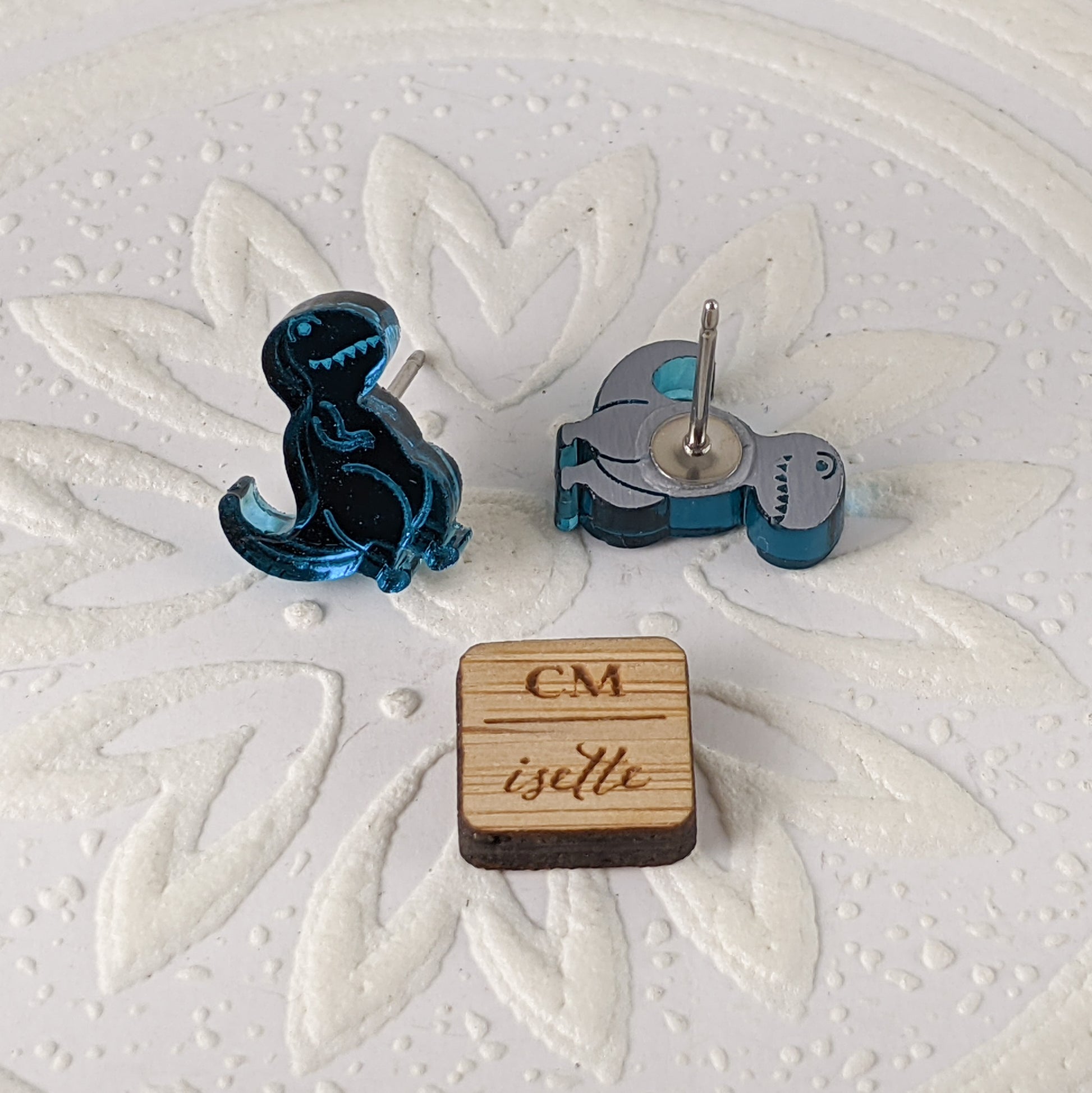 Cartoon Tyrannosaurus Rex stud earrings in teal mirrored acrylic.  They are photographed with one standing, one face down, next to a 1 cm wooden square for size comparison. By Isette.