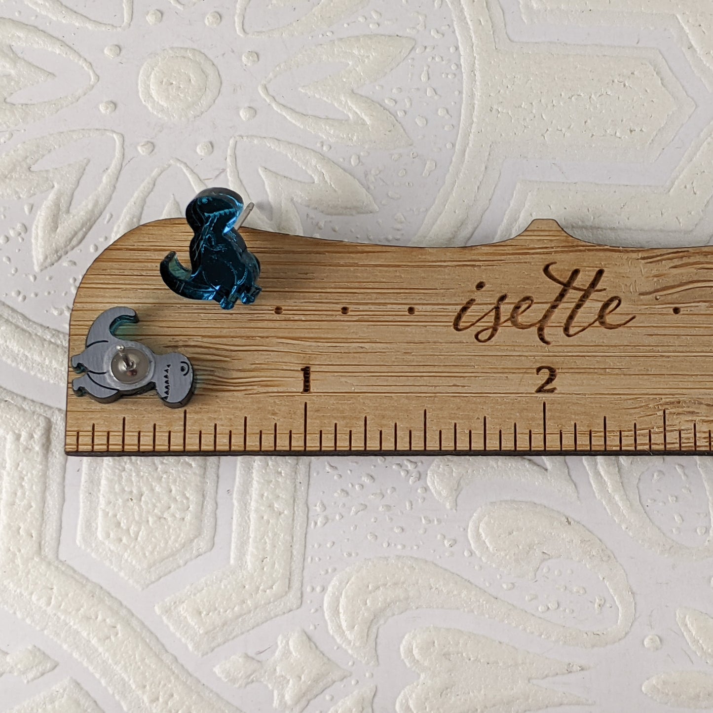 oon Tyrannosaurus Rex stud earrings in teal mirrored acrylic. They are photographed with one standing, one face down, on a wooden ruler, and measure .5 inches.  By Isette.