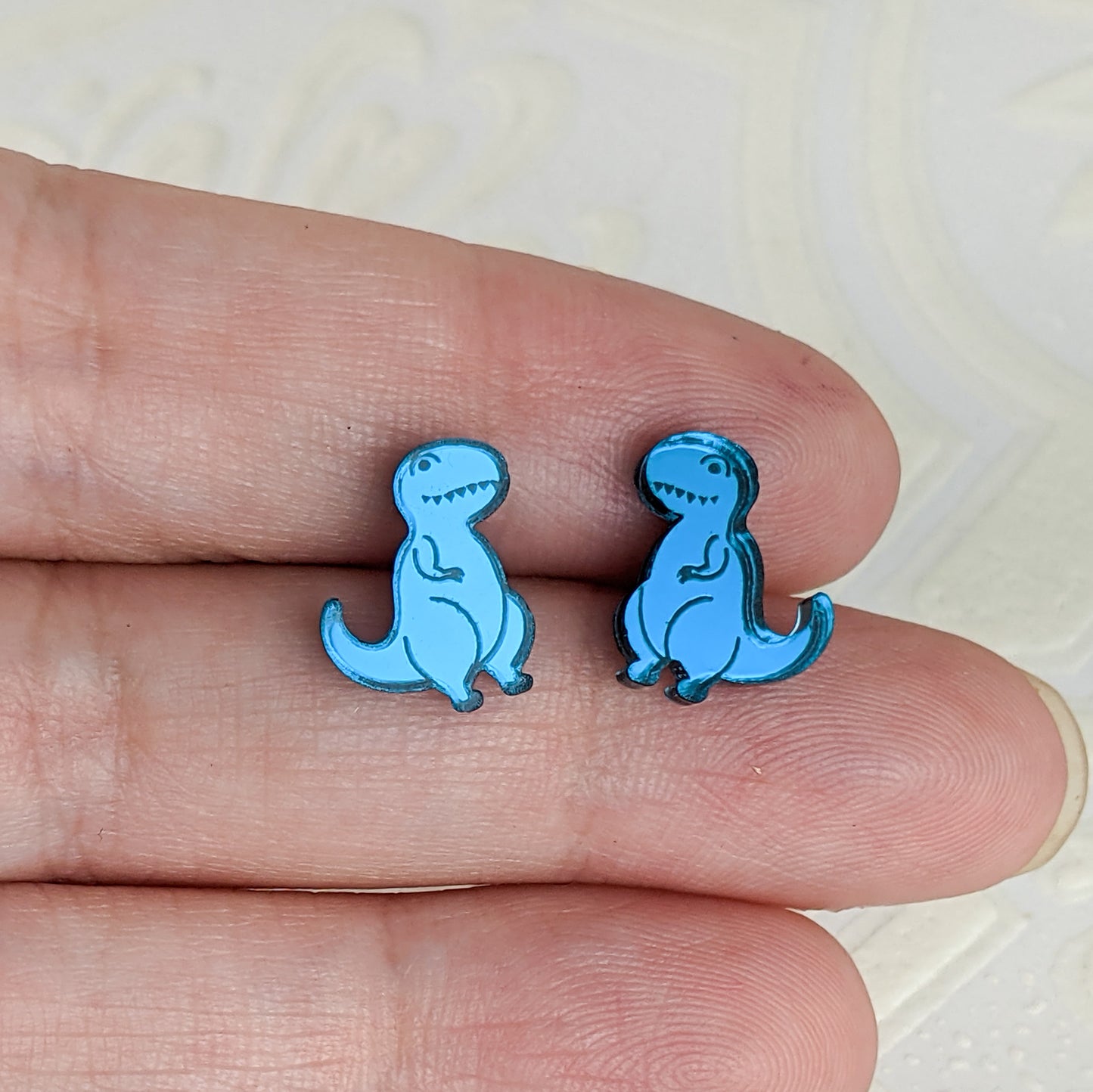 Cartoon Tyrannosaurus Rex stud earrings in teal mirrored acrylic.  The dinosaurs face towards each other and are being held between fingers on an open hand to show they are roughly the height of a finger.  By Isette.