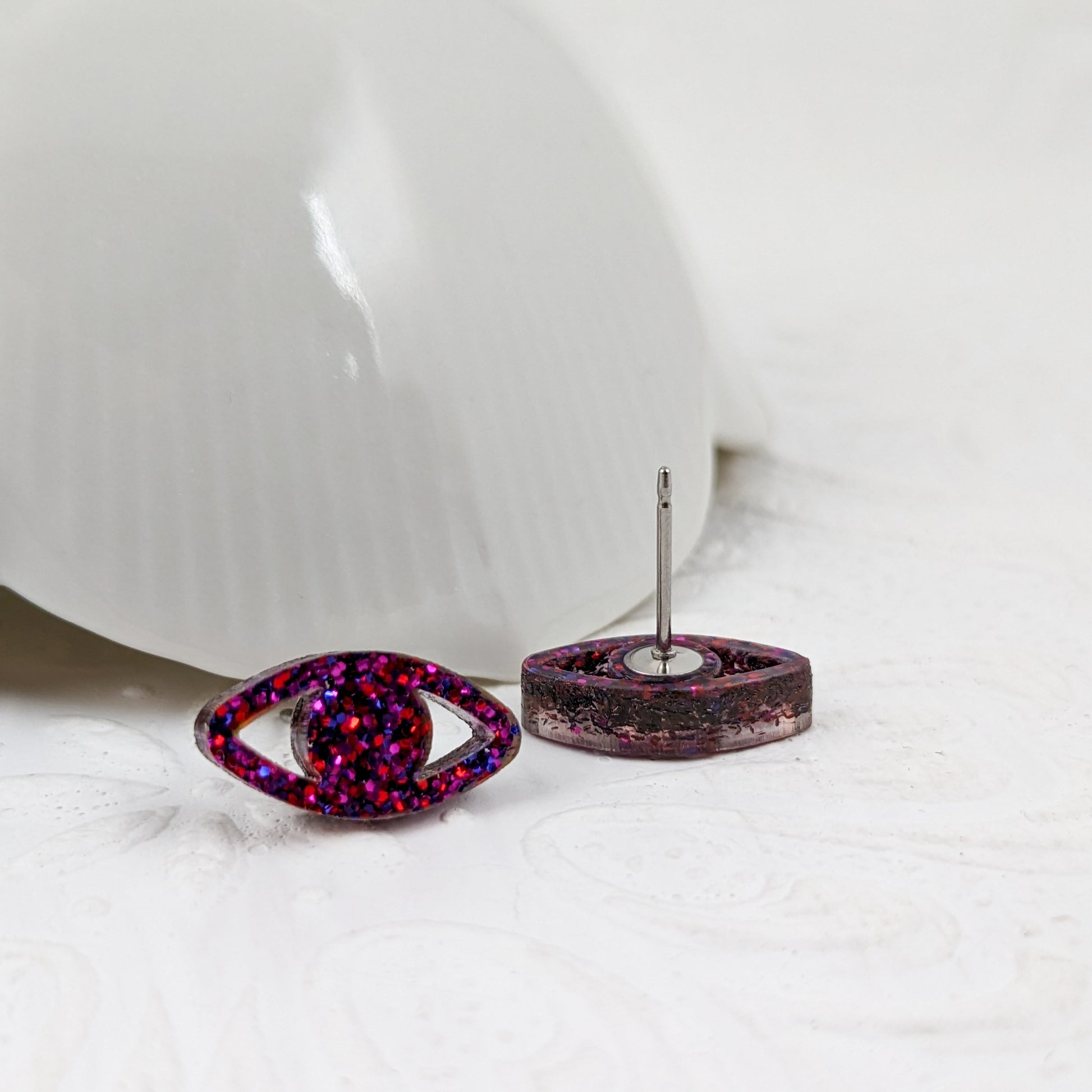 Peepers - Eye shaped stud earrings made with ruby sparkle acrylic and stainless steel posts in front of a vintage white dish