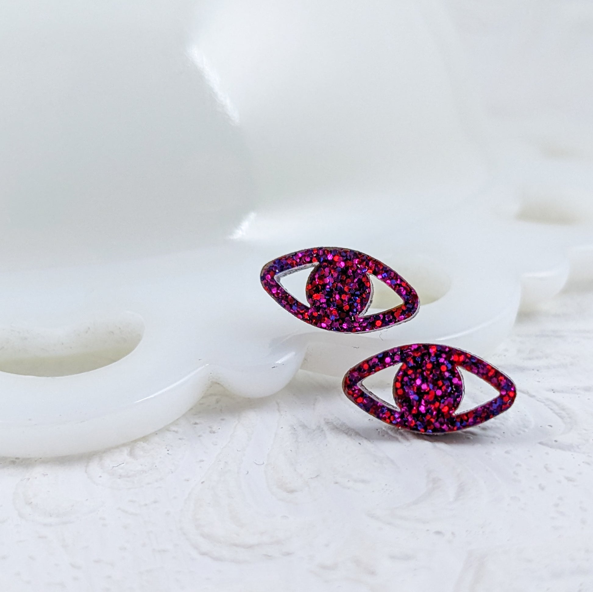 Peepers - Eye shaped stud earrings made with ruby sparkle acrylic and stainless steel posts in front of vintage white milkglass