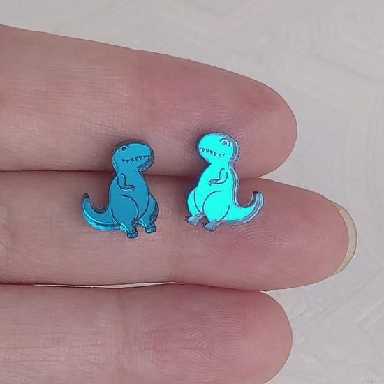 Video of Cartoon Tyrannosaurus Rex stud earrings in teal mirrored acrylic being held between two fingers.  They are being slowly moved to show how the light reflects off the mirrored surface.  By Isette.