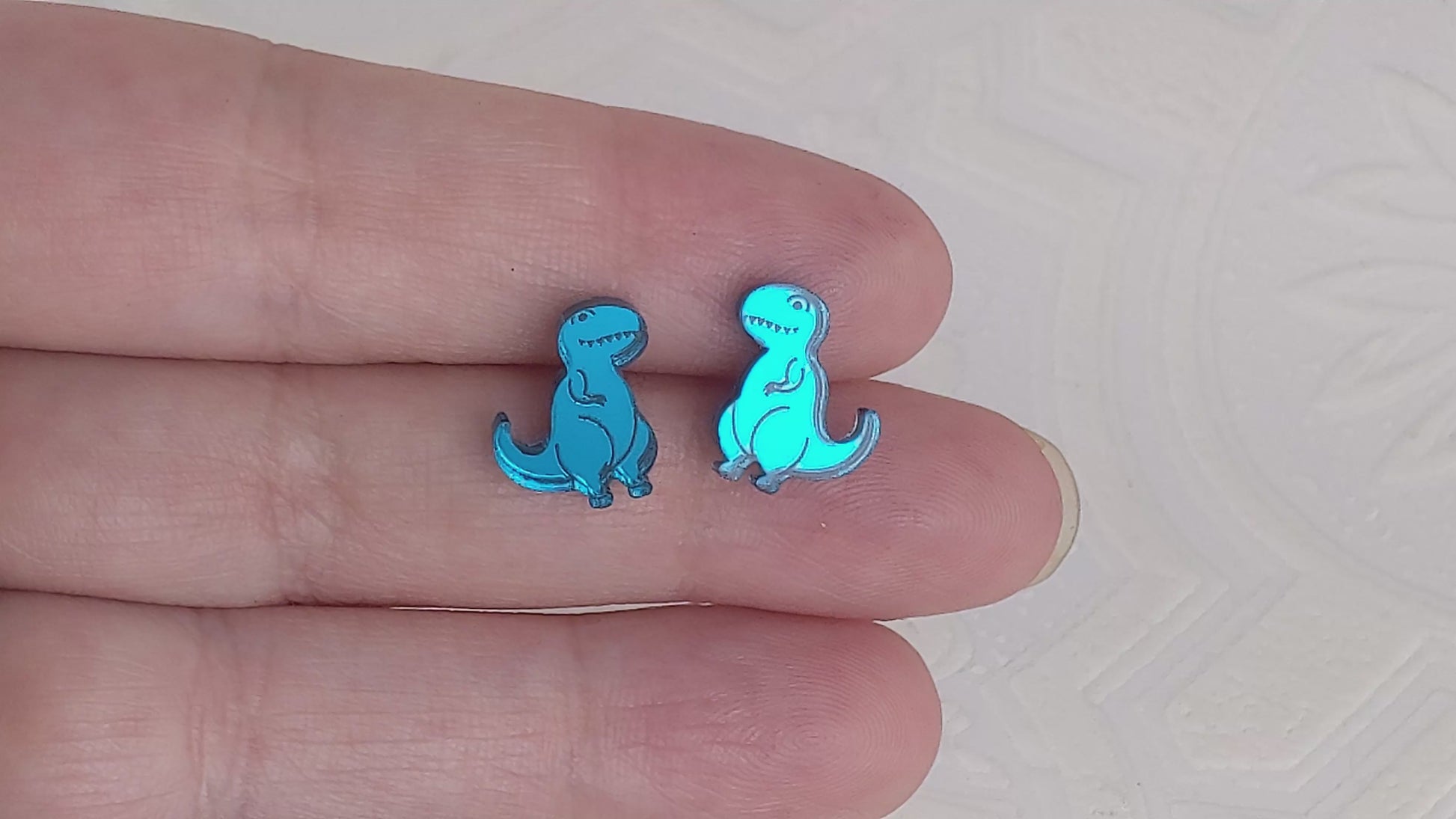 Video of Cartoon Tyrannosaurus Rex stud earrings in teal mirrored acrylic being held between two fingers.  They are being slowly moved to show how the light reflects off the mirrored surface.  By Isette.